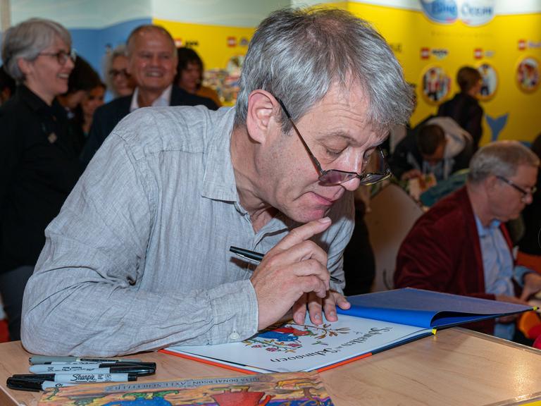 Der Illustrator Axel Scheffler signiert auf der Frankfurter Buchmesse Bücher. Er trägt ein graues Hemd, hat einen Stift in der Hand und ist über ein Buch gebeugt.