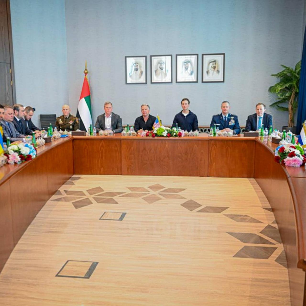 An einem u-förmigen Tisch sitzen mehrere Männer. Ukrainische und russische Flaggen stehen zwischen ihnen auf dem Tisch.