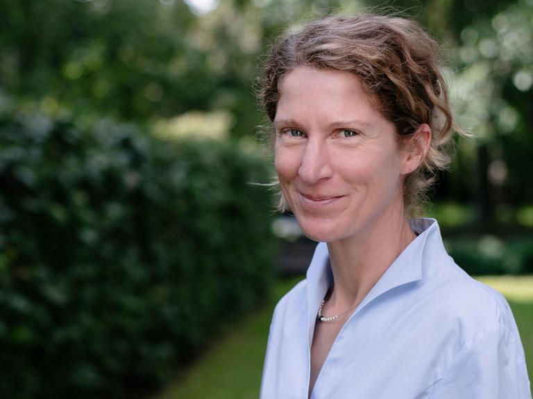 Eine Frau mit dunkelblonden, hochgesteckten Haaren und hellblauer Bluse schaut lächelnd.