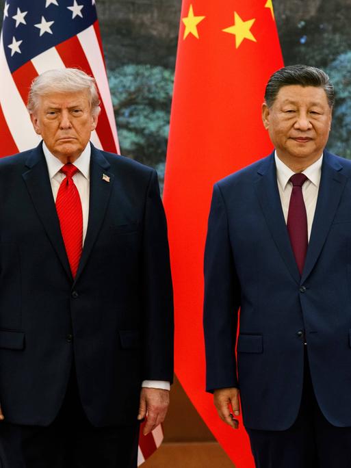 Der Präsident der USA Donald Trump mit dem chinesischen Präsidenten Xi Jinping. Bei einem Treffen stehen sie nebeneinander. Beide tragen blaue Anzüge und rote Krawatten. Sie schauen in Richtung der Kamera. Im Hintergrund die chinesische und die US-Flagge.