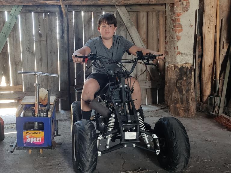 Ein Junge sitzt auf einem Quad. Daneben steht das von ihm aus einer Getränkekiste und einem Roller gebaute Gefährt.