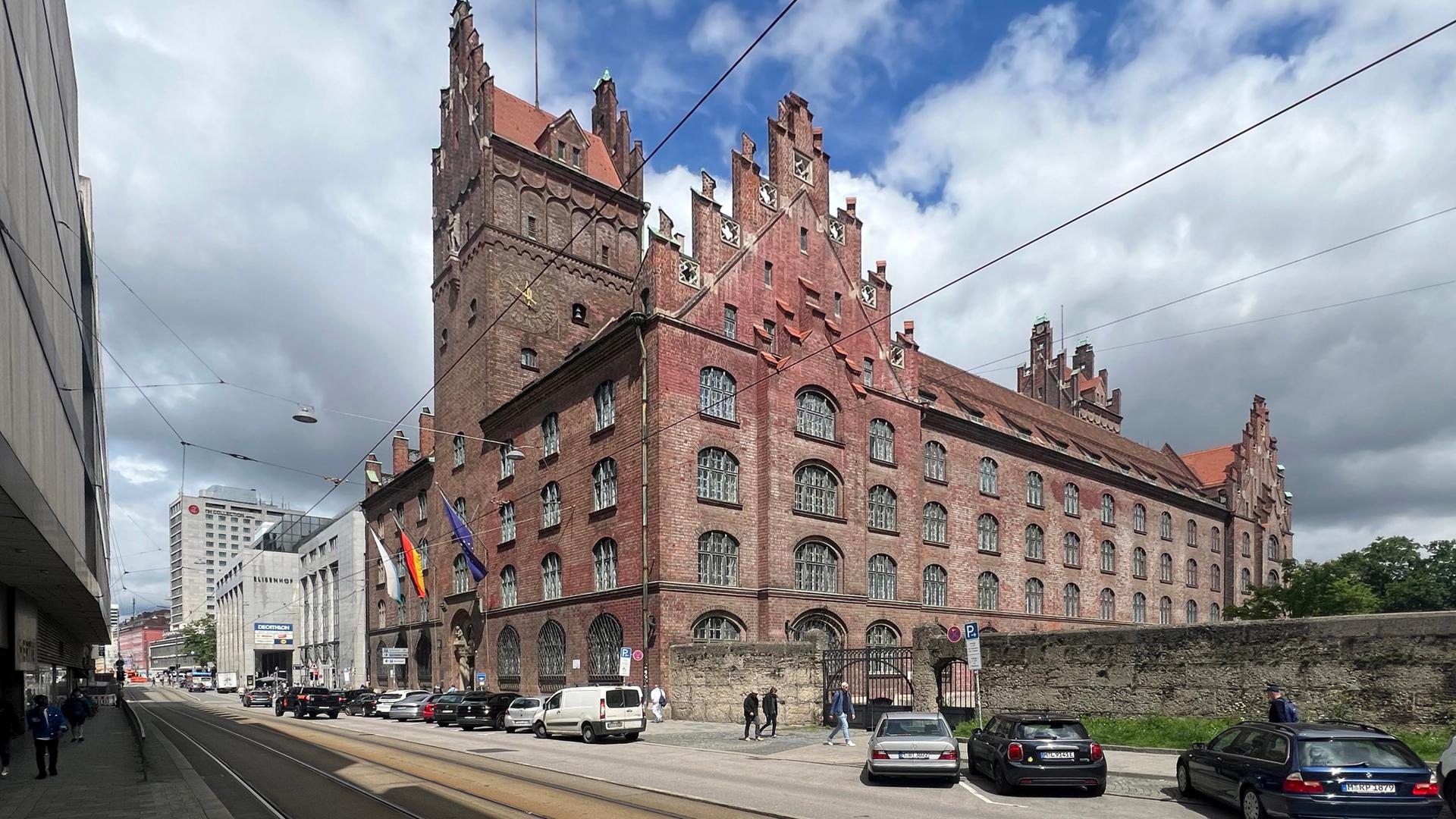 Blick auf das große burgähnliche Backsteingebäude unter einem Wolkenhimmel. Davor parken Autos. Auf den Bürgersteigen gehen Menschen.