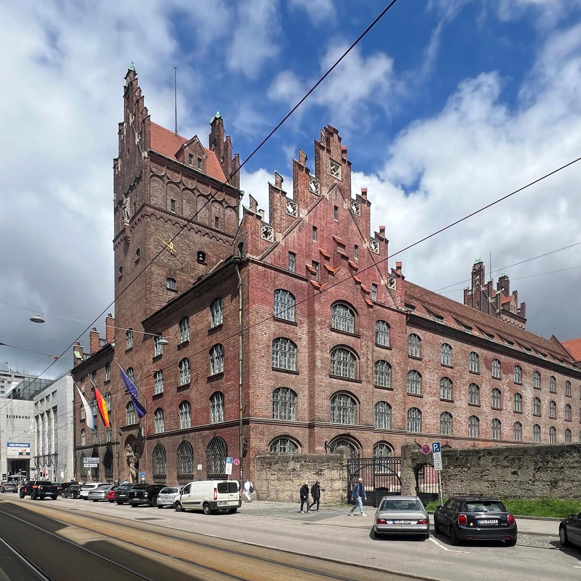 Blick auf das große burgähnliche Backsteingebäude unter einem Wolkenhimmel. Davor parken Autos. Auf den Bürgersteigen gehen Menschen.