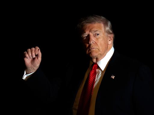 Donald Trump im Anzug und mit roter Krawatte wird in Dunkelheit vom Blitzlicht einer Kamera beleuchtet, während er mit der rechten Hand gestikuliert