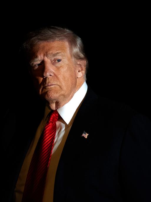 Donald Trump im Anzug und mit roter Krawatte wird in Dunkelheit vom Blitzlicht einer Kamera beleuchtet, während er mit der rechten Hand gestikuliert