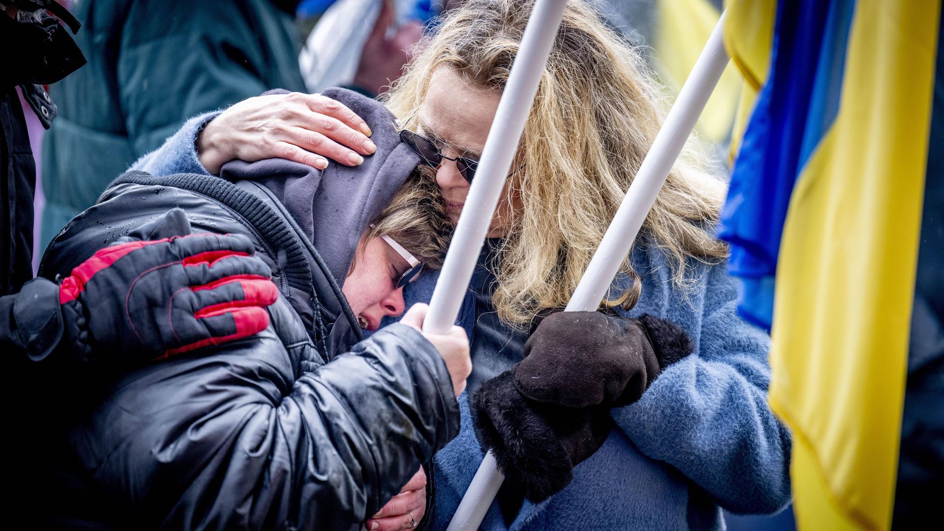 Zwei Frauen liegen sich in den Armen. Sie trauern. Sie tragen Fahnen von der Ukraine.