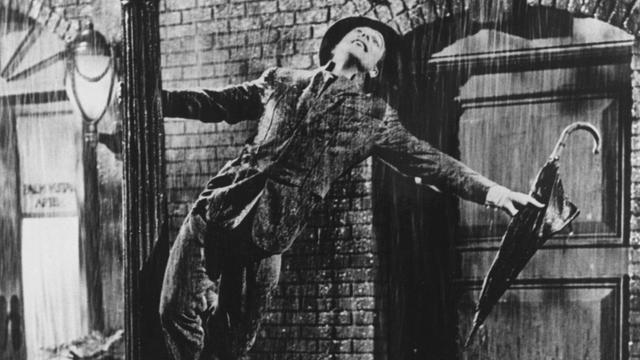 Eine Szene aus dem Film "Singin' in the Rain" zeigt den Schauspieler Gene Kelly, der mit einem nicht ausgeklappten Regenschirm in der Hand im Regen auf dem Sockel einer Straßenlaterne steht und in den Regen hinauf schaut