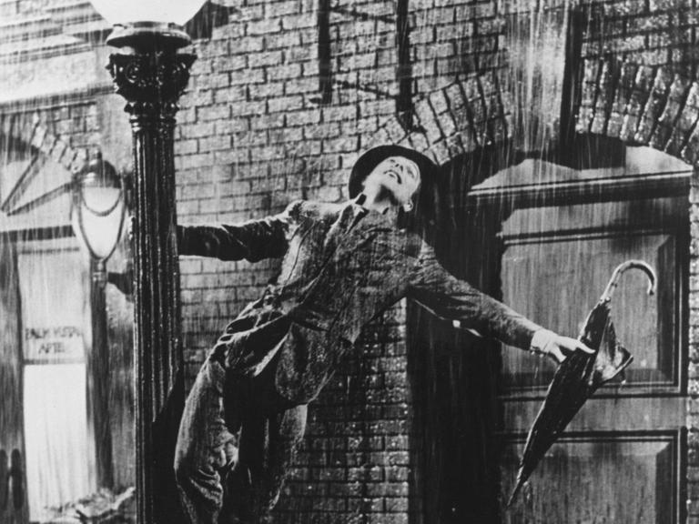 Eine Szene aus dem Film "Singin' in the Rain" zeigt den Schauspieler Gene Kelly, der mit einem nicht ausgeklappten Regenschirm in der Hand im Regen auf dem Sockel einer Straßenlaterne steht und in den Regen hinauf schaut