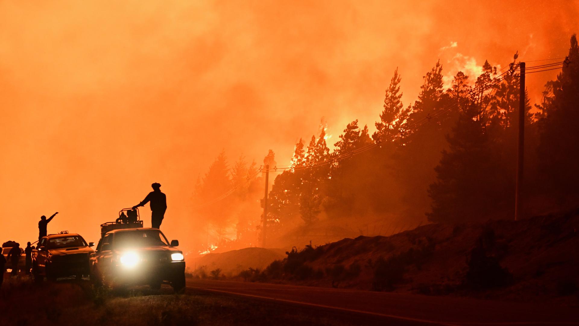 Feuerwehrleute stehen auf einem Lastwagen, während im Hintergrund Waldbrände lodern. 