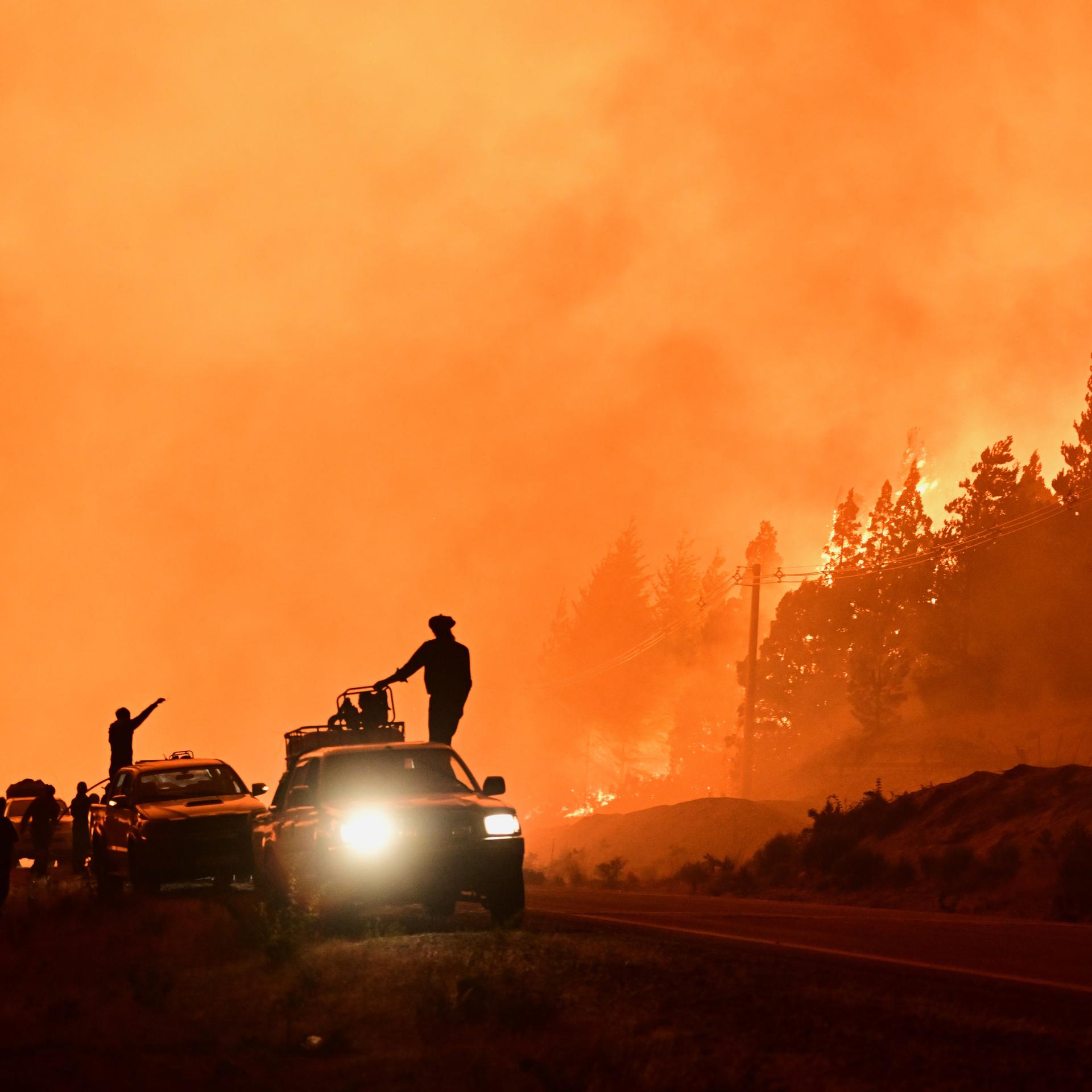 Feuerwehrleute stehen auf einem Lastwagen, während im Hintergrund Waldbrände lodern. 