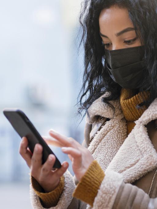 Eine Frau mit einer Maske vor Nase und Muns steht auf einer Straße und hält dabei ein Handy in der Hand.