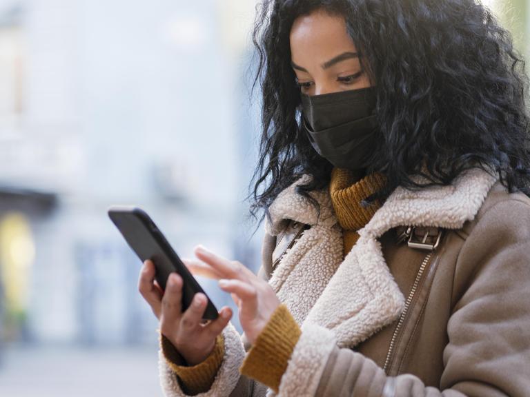 Eine Frau mit einer Maske vor Nase und Muns steht auf einer Straße und hält dabei ein Handy in der Hand.