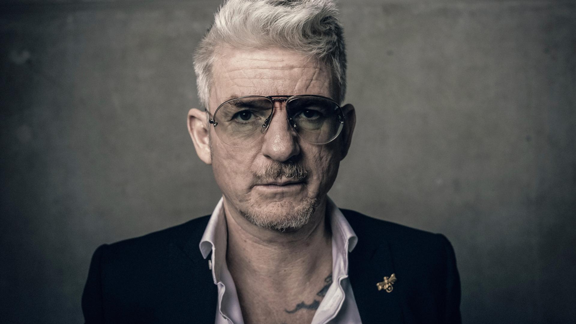 Porträt von Heinz Strunk. Er trägt eine Pilotenbrille und ein Sakko mit weit geöffnetem Hemdkragen. Porträt von Heinz Strunk. Er trägt eine Pilotenbrille und ein Sakko mit weit geöffnetem Hemdkragen.