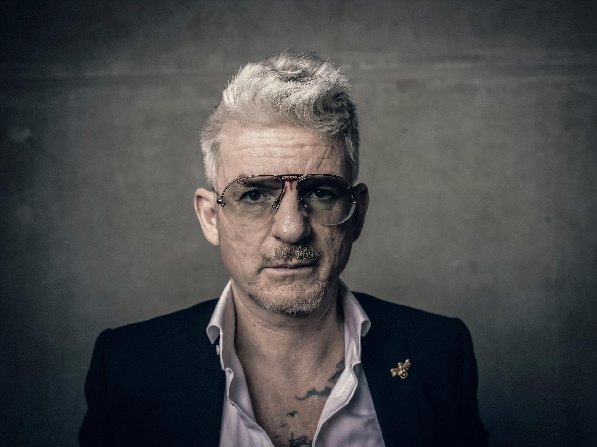 Porträt von Heinz Strunk. Er trägt eine Pilotenbrille und ein Sakko mit weit geöffnetem Hemdkragen. Porträt von Heinz Strunk. Er trägt eine Pilotenbrille und ein Sakko mit weit geöffnetem Hemdkragen.