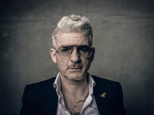 Porträt von Heinz Strunk. Er trägt eine Pilotenbrille und ein Sakko mit weit geöffnetem Hemdkragen. Porträt von Heinz Strunk. Er trägt eine Pilotenbrille und ein Sakko mit weit geöffnetem Hemdkragen.