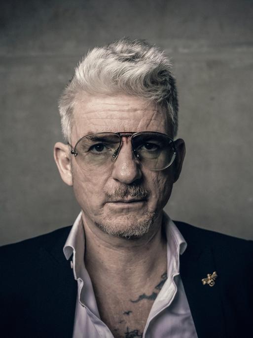 Porträt von Heinz Strunk. Er trägt eine Pilotenbrille und ein Sakko mit weit geöffnetem Hemdkragen. Porträt von Heinz Strunk. Er trägt eine Pilotenbrille und ein Sakko mit weit geöffnetem Hemdkragen.