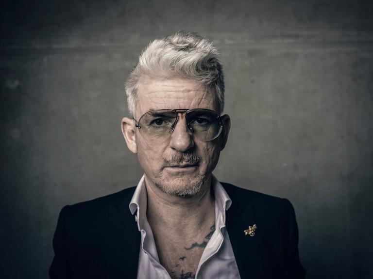 Porträt von Heinz Strunk. Er trägt eine Pilotenbrille und ein Sakko mit weit geöffnetem Hemdkragen. Porträt von Heinz Strunk. Er trägt eine Pilotenbrille und ein Sakko mit weit geöffnetem Hemdkragen.