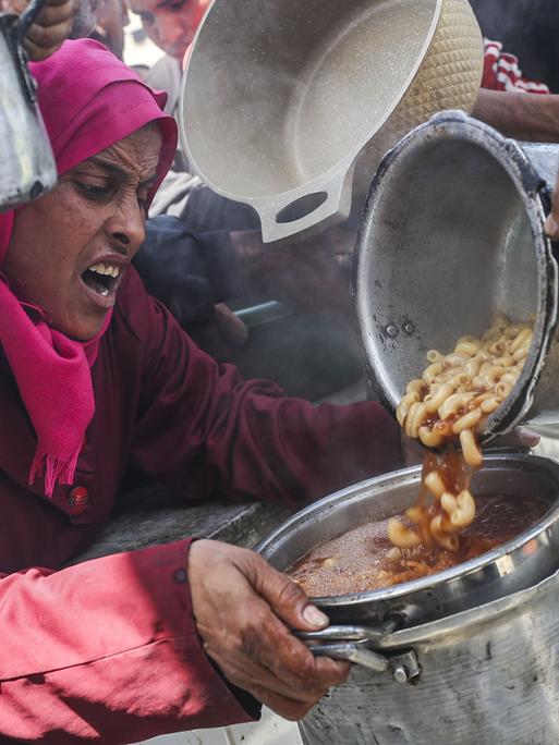 Eine Frau mit verzweifeltem Gesichtsausdruck bekommt Essen in ihren Topf gefüllt. Um sie herum drängen sich Menschen mit leeren Töpfen.