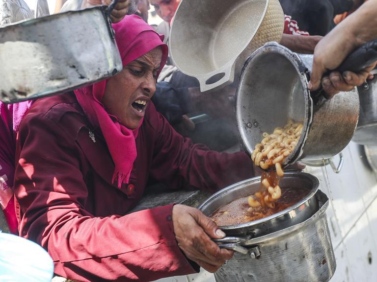 Eine Frau mit verzweifeltem Gesichtsausdruck bekommt Essen in ihren Topf gefüllt. Um sie herum drängen sich Menschen mit leeren Töpfen.