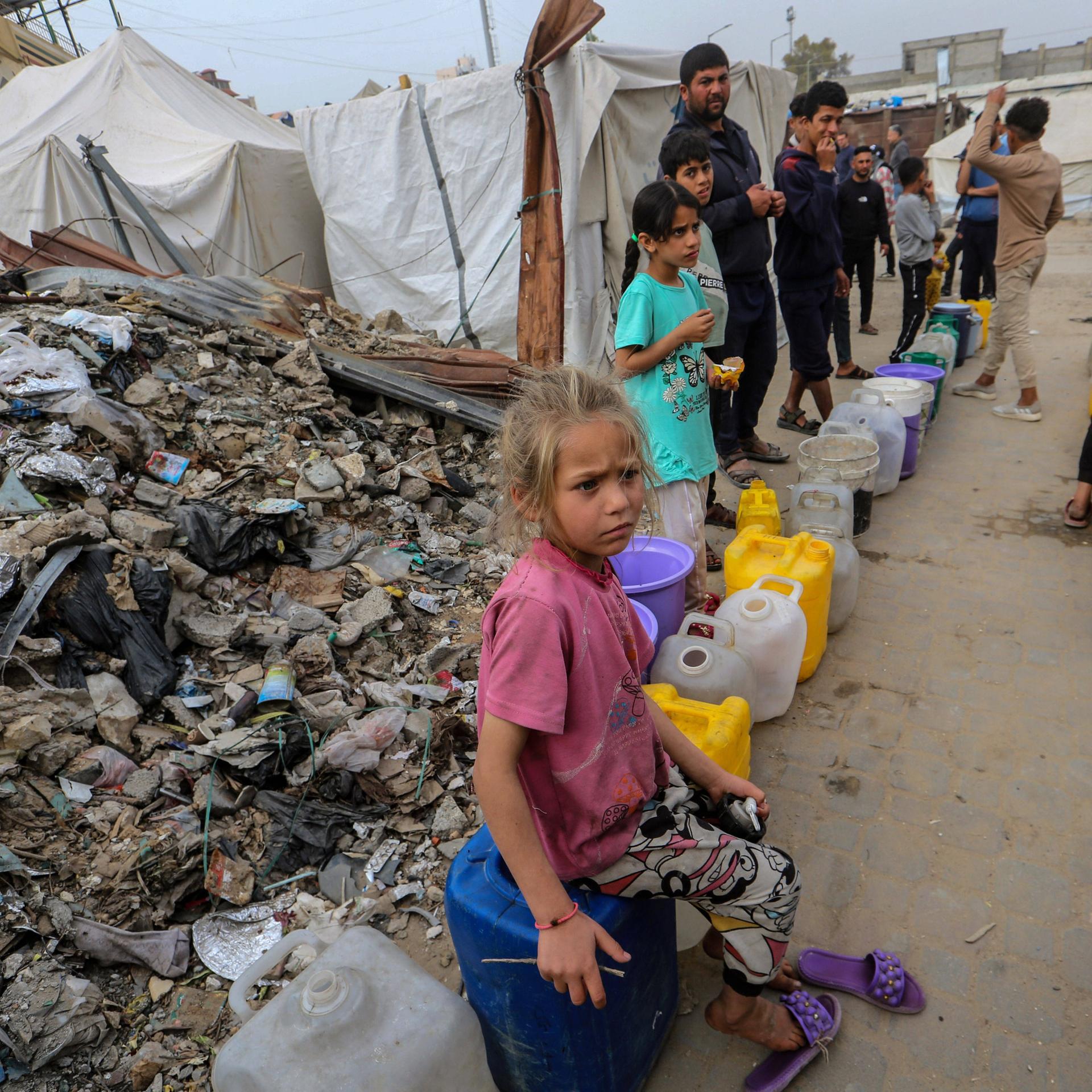 Am Rande eines Trümmerfeldes warten Kinder in Gaza mit bunten Plastikkanistern auf die Ausgabe von frischem Trinkwasser.