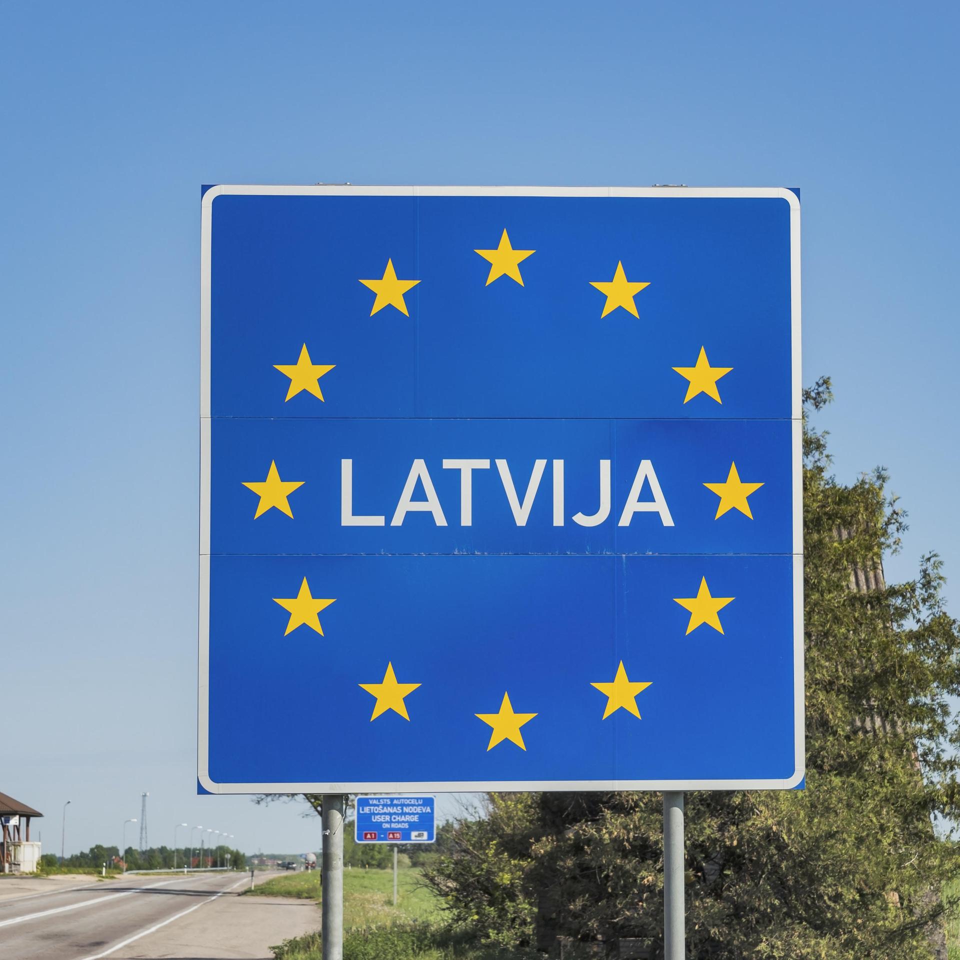 Das große blaue Schild mit der Aufschrift "Latvija" und dem europäischen Sternenkreis nimmt den Großteil des Bildes ein. Am unteren Rand der Grenzübergang mit einem Gebäude, dahinter blauer Himmel. 