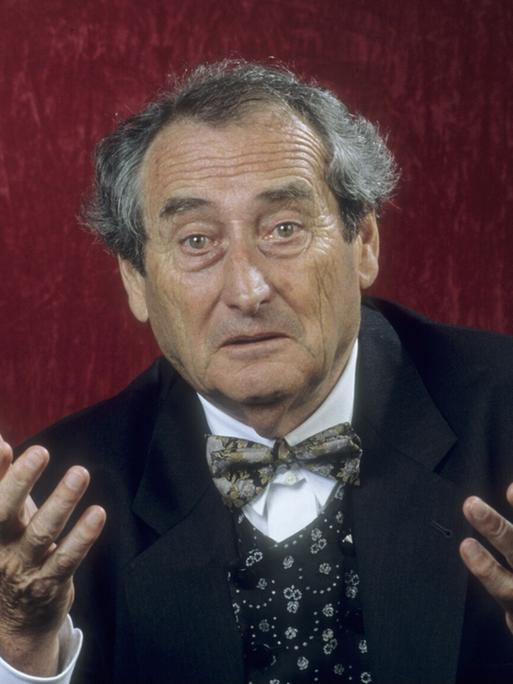 Schulheiß mit dunklem Anzug und Fliege steht vor einer roten Wand. Er hält mit fragendem Blick beide Hände hoch. 