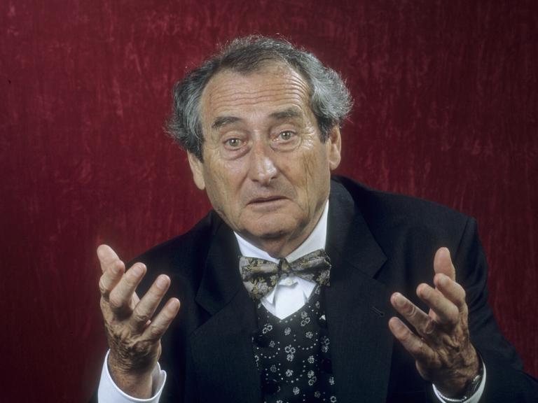 Schulheiß mit dunklem Anzug und Fliege steht vor einer roten Wand. Er hält mit fragendem Blick beide Hände hoch. 