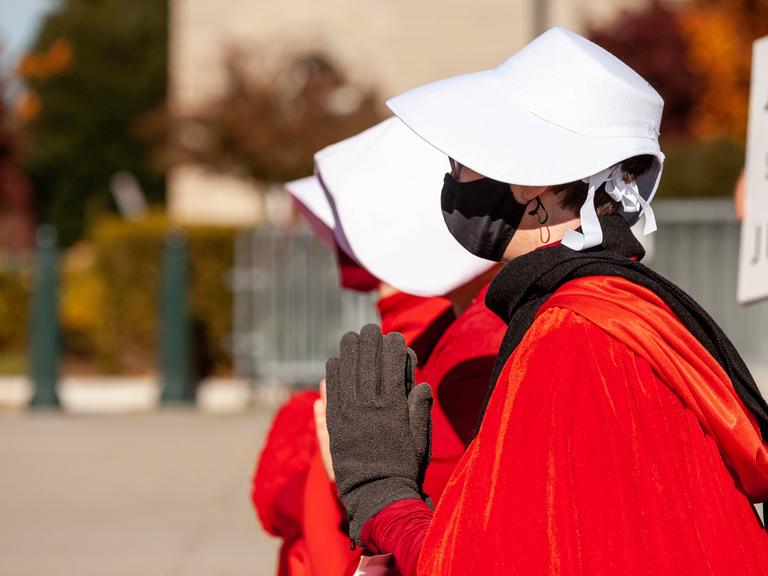 Frauen, gekleidet mit einem roten Umhang und einer weißen Haube, stehen auf einem Platz und halten die Hände zusammengedrückt vor den Oberkörper.