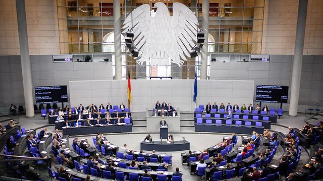 Blick in den großen Raum von dem Bundestag. Abgeordnete sitzen auf blauen Stühlen.