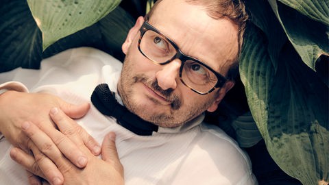 Milan Peschenl liegt zwischen den Blättern einer Topfpflanze, trägt ein weisses Hemd und eine schwarze Fliege und schaut schelmisch zur Seite. Milan Peschenl liegt zwischen den Blättern einer Topfpflanze, trägt ein weisses Hemd und eine schwarze Fliege und schaut schelmisch zur Seite.