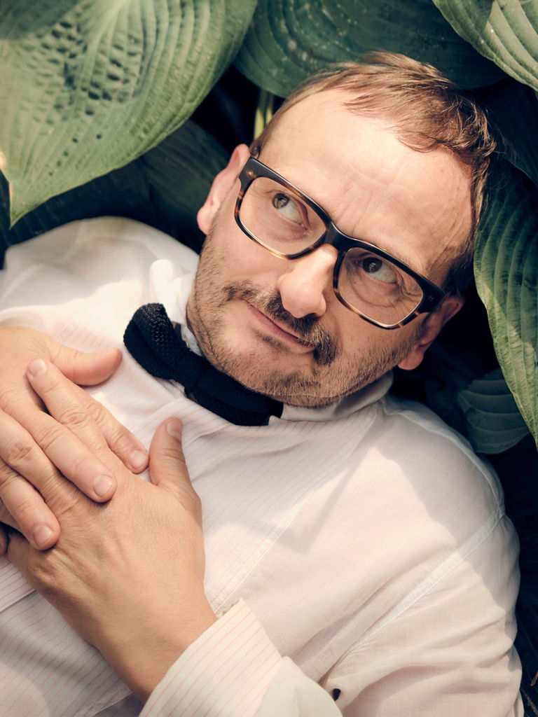 Milan Peschenl liegt zwischen den Blättern einer Topfpflanze, trägt ein weisses Hemd und eine schwarze Fliege und schaut schelmisch zur Seite.