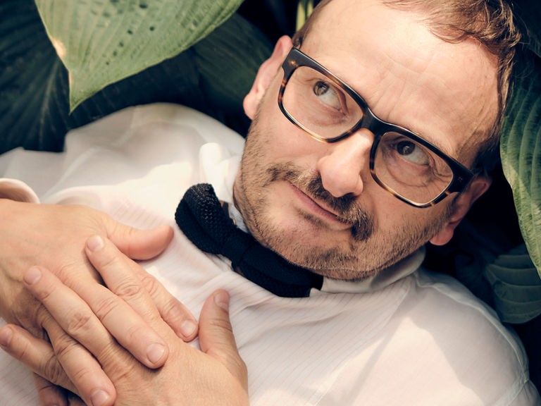 Milan Peschenl liegt zwischen den Blättern einer Topfpflanze, trägt ein weisses Hemd und eine schwarze Fliege und schaut schelmisch zur Seite. Milan Peschenl liegt zwischen den Blättern einer Topfpflanze, trägt ein weisses Hemd und eine schwarze Fliege und schaut schelmisch zur Seite.