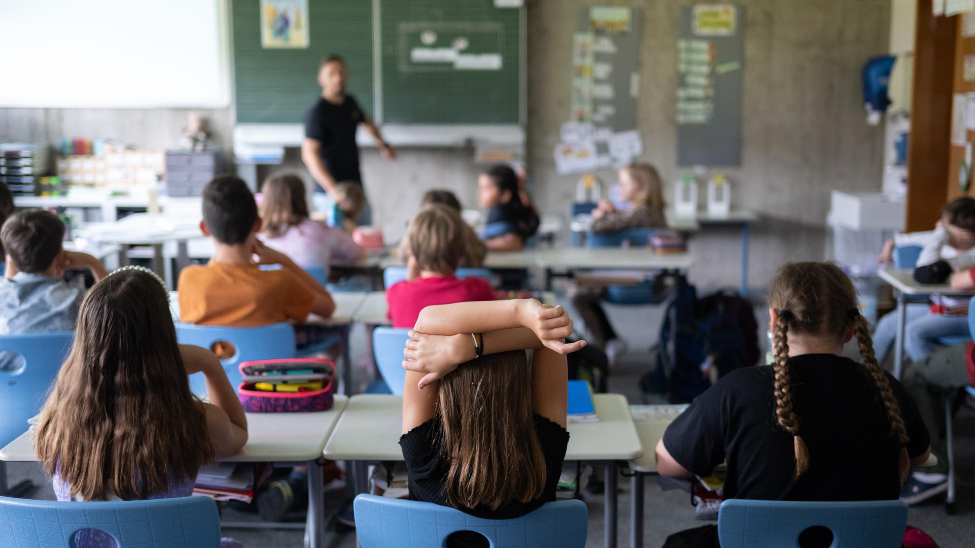 Schülerinnen und Schüler einer vierten Klasse sitzen während ihrer Stunde im Klassenzimmer. Im Hintergrund sieht man unscharf einen Lehrer an der Tafel.