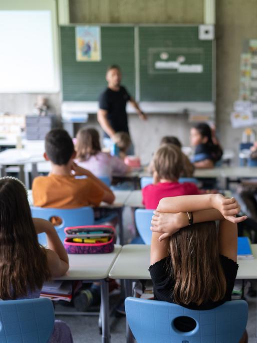 Schülerinnen und Schüler einer vierten Klasse sitzen während ihrer Stunde im Klassenzimmer. Im Hintergrund sieht man unscharf einen Lehrer an der Tafel.