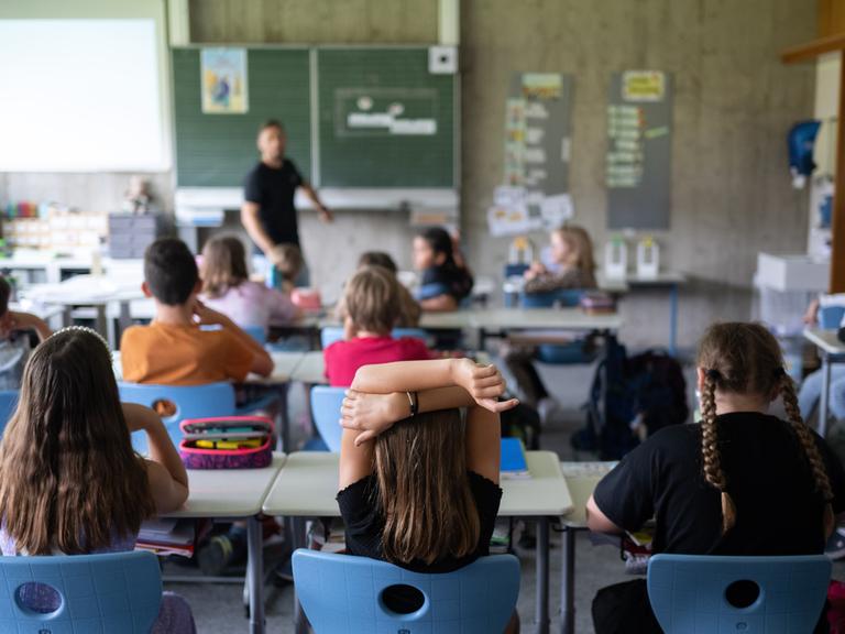 Schülerinnen und Schüler einer vierten Klasse sitzen während ihrer Stunde im Klassenzimmer. Im Hintergrund sieht man unscharf einen Lehrer an der Tafel.