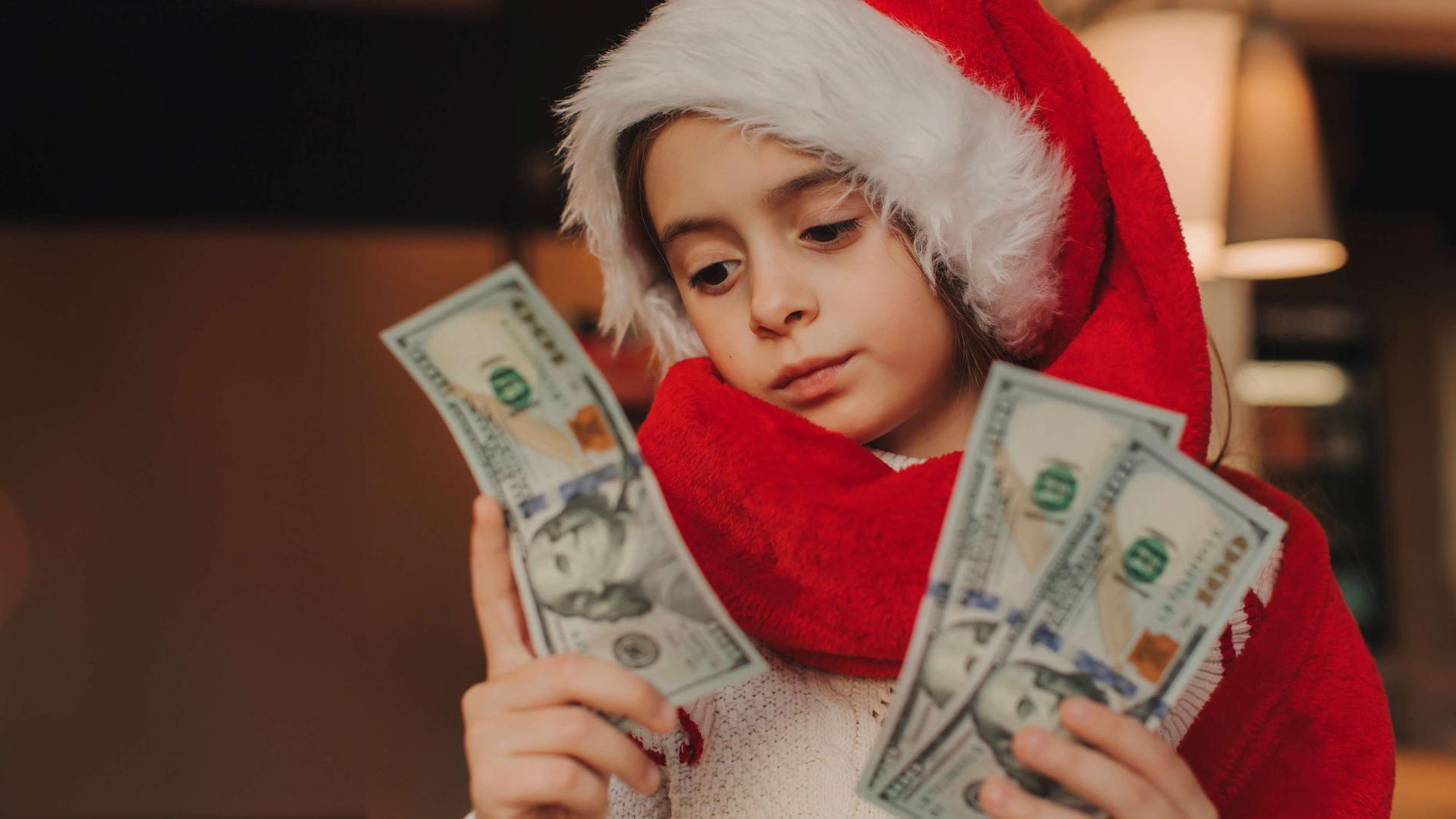 Ein Kind im Schulalter trägt eine Weihnachtsmann-Mütze und Weihnachtsmann-Mantel und blickt auf drei Dollar-Scheine in der Hand.