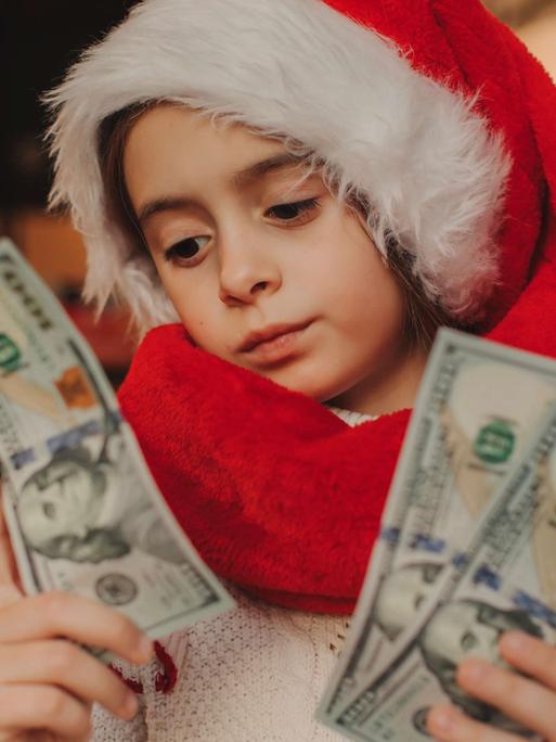 Ein Kind im Schulalter trägt eine Weihnachtsmann-Mütze und Weihnachtsmann-Mantel und blickt auf drei Dollar-Scheine in der Hand.