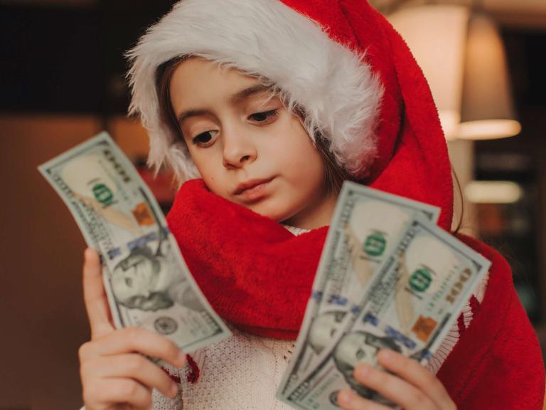 Ein Kind im Schulalter trägt eine Weihnachtsmann-Mütze und Weihnachtsmann-Mantel und blickt auf drei Dollar-Scheine in der Hand.