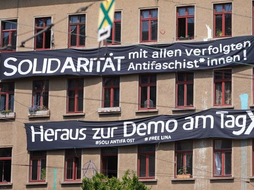 Transparente mit der Aufschrift "Solidarität mit allen verfolgten Antifaschist*innen!" und "Heraus zur Demo am Tag X" hängen an der Fassade eines Hauses.