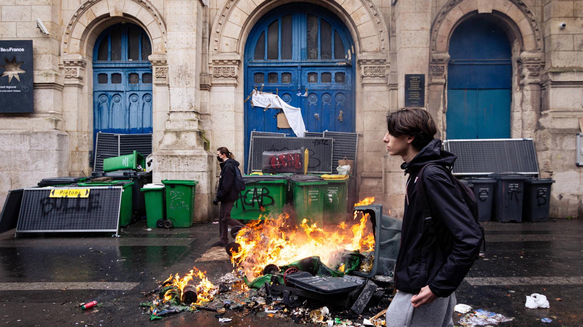 Junge Menschen gehen in Paris an brennenden Mülleimern vorbei. Vor der Tür des Voltaire-Gymnasiums während einer Demonstration am 1. Februar 2024.