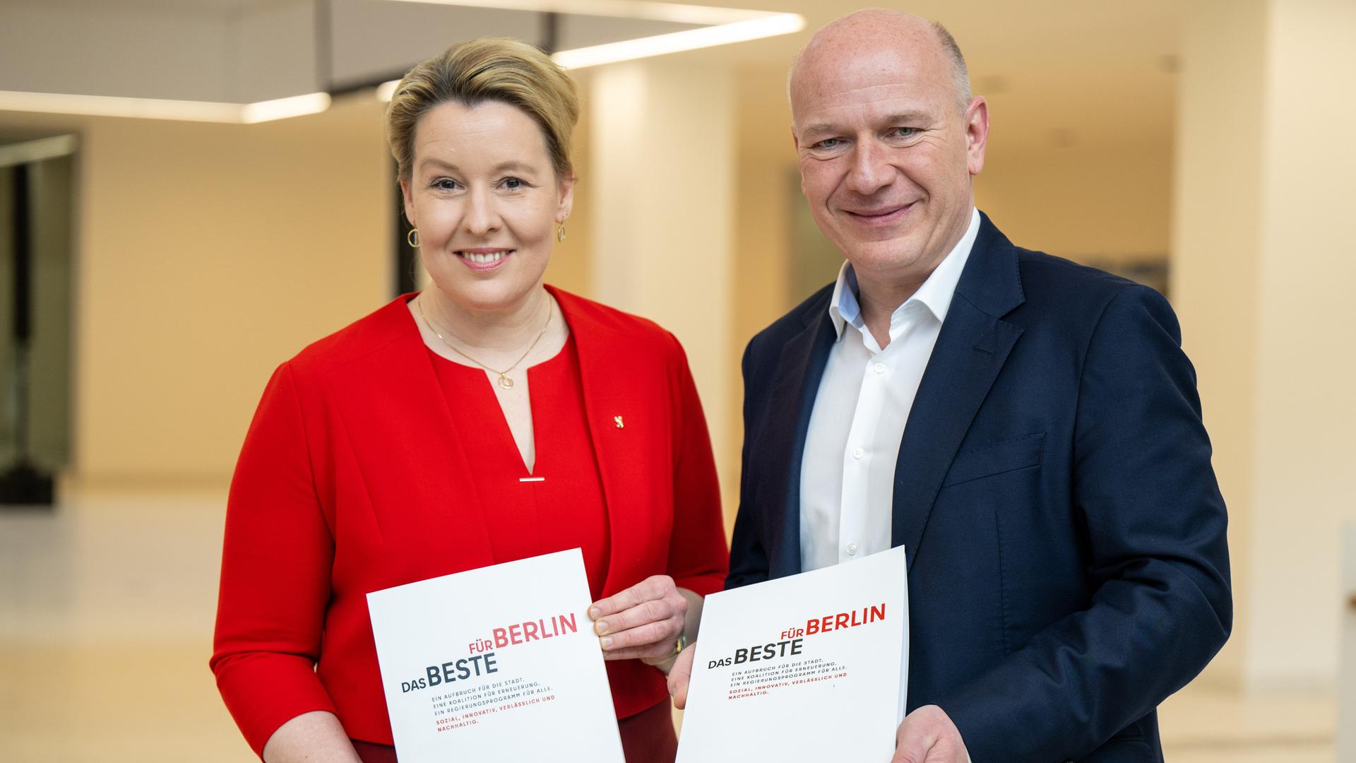 Franziska Giffey (links), in einem roten Kleid, und Kai Wegner, in einem dunklen Anzug, halten je ein Exemplar ihres Koalitionsvertrags in Händen, Aufschrift: "Das Beste für Berlin".