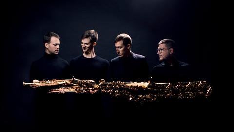 Vier junge Männer halten ihre unterschiedlich großen Saxophone quer, sodass er schient, als ob die Saxophone ein riesiges, goldenes Instrument ergeben.