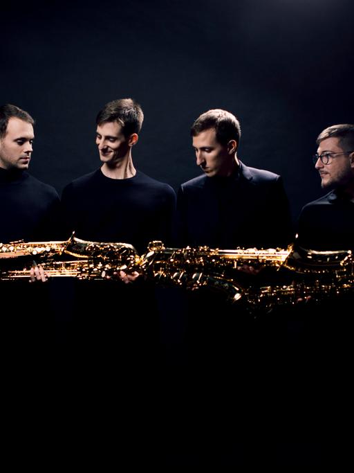 Vier junge Männer halten ihre unterschiedlich großen Saxophone quer, sodass er schient, als ob die Saxophone ein riesiges, goldenes Instrument ergeben.