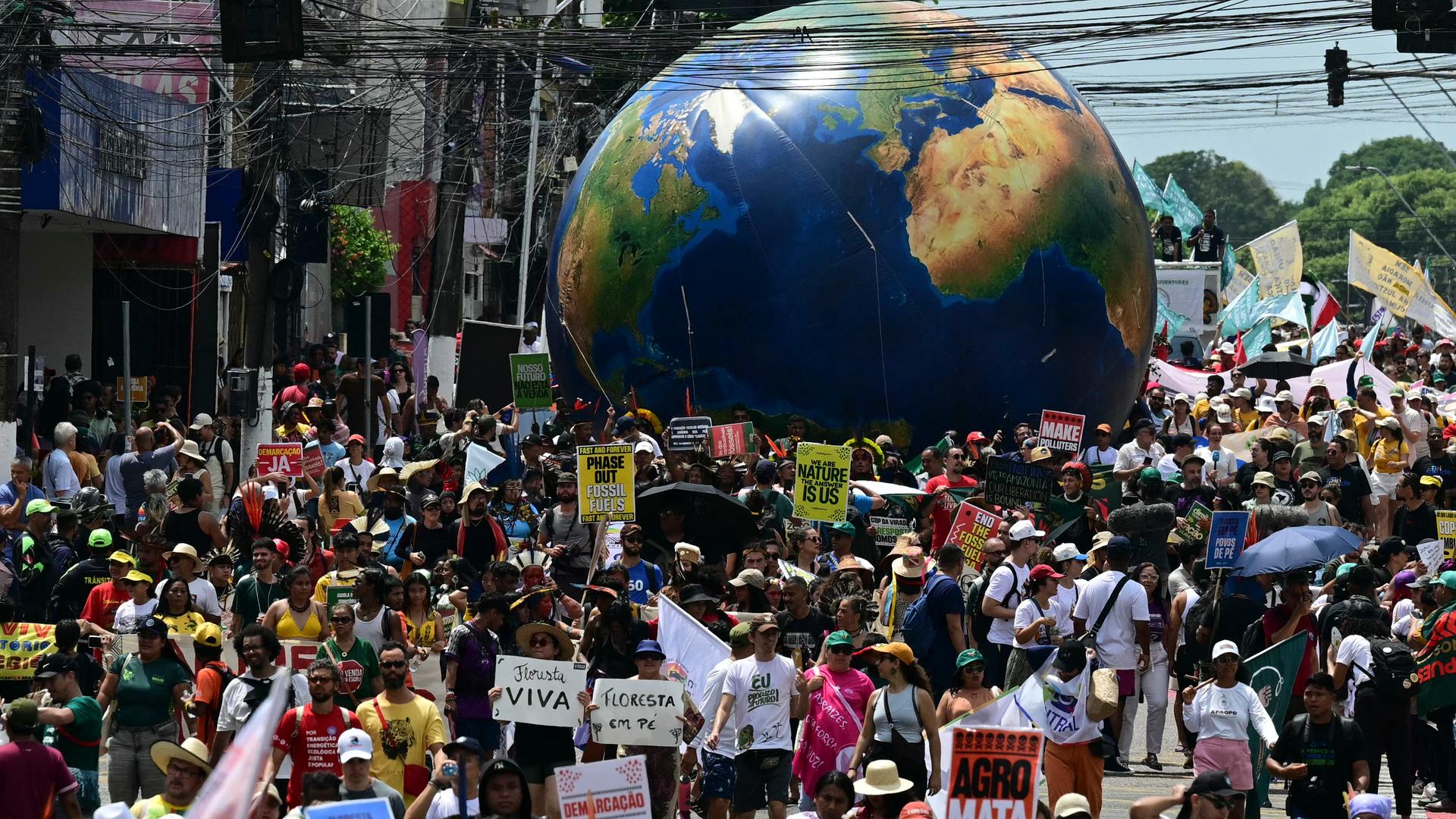 Tausende Menschen nehmen in Belem am sogenannten "Marsch fürs Klima" teil. Zu sehen sind viele Menschen auf einer Straße, einige halten Transparente hoch; im Hintergrund ist eine riesige Weltkugel zu sehen. 