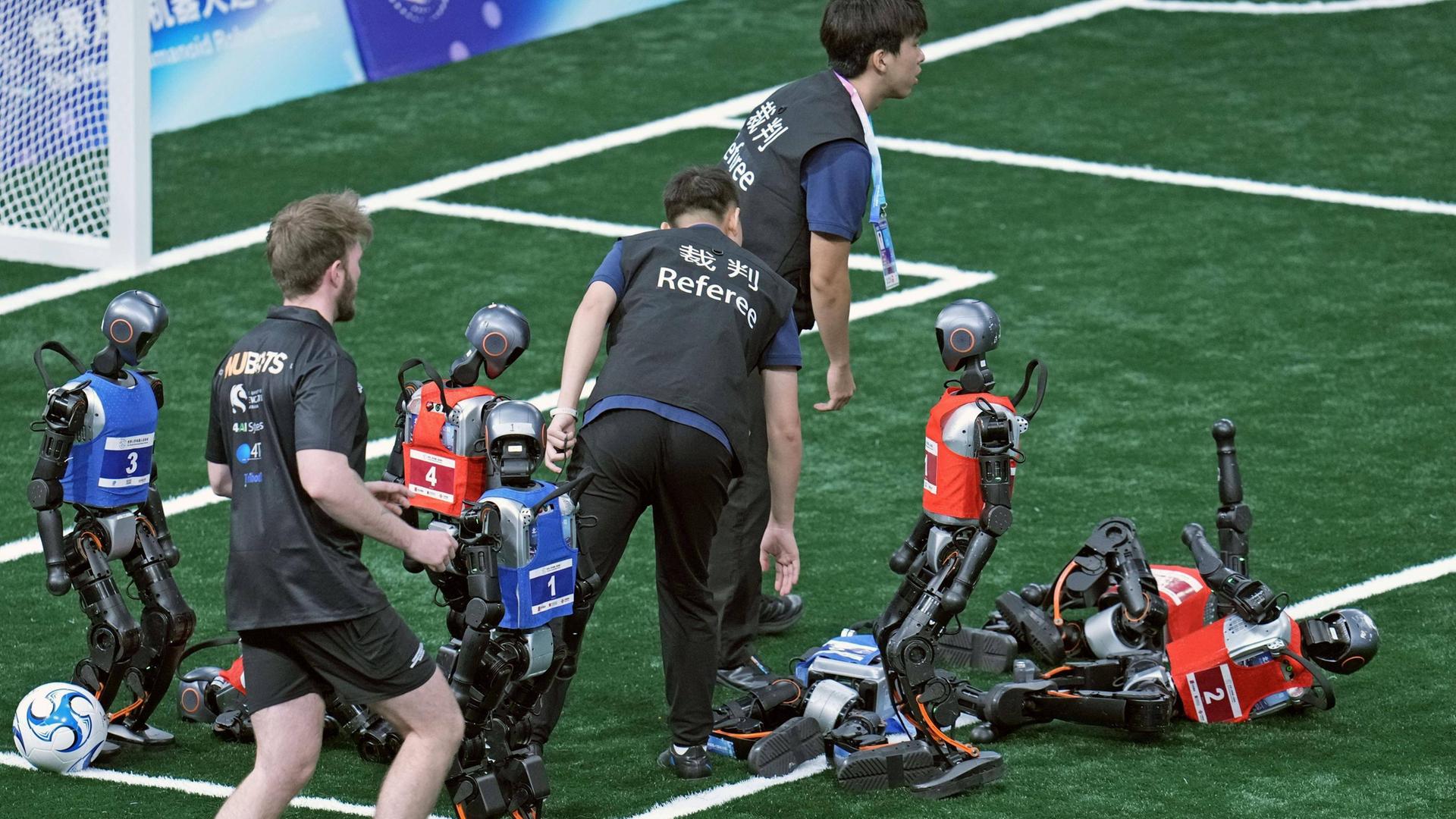 Ein Fußballspiel von Robotern. Die Roboter sind so groß wie Kinder. Drei Roboter liegen am Boden. Dazwischen stehen Menschen. 