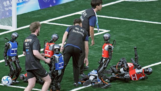 Ein Fußballspiel von Robotern. Die Roboter sind so groß wie Kinder. Drei Roboter liegen am Boden. Dazwischen stehen Menschen. 