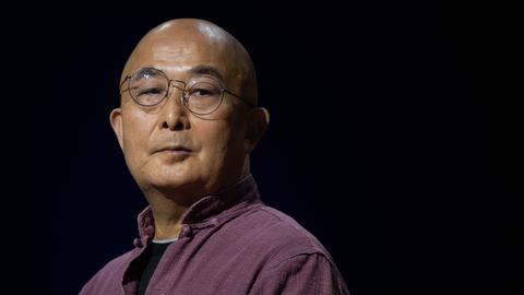 Der chinesische Schriftsteller Liao Yiwu blickt im lila Hemd mit hochgestelltem Kragen, mit Nickelbrille und kahlgeschorenem Kopf über seine Schulter.