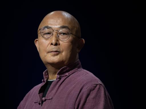 Der chinesische Schriftsteller Liao Yiwu blickt im lila Hemd mit hochgestelltem Kragen, mit Nickelbrille und kahlgeschorenem Kopf über seine Schulter.