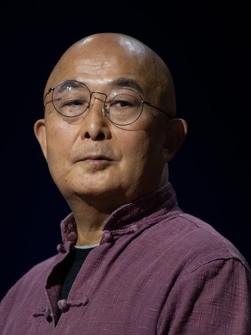 Der chinesische Schriftsteller Liao Yiwu blickt im lila Hemd mit hochgestelltem Kragen, mit Nickelbrille und kahlgeschorenem Kopf über seine Schulter.