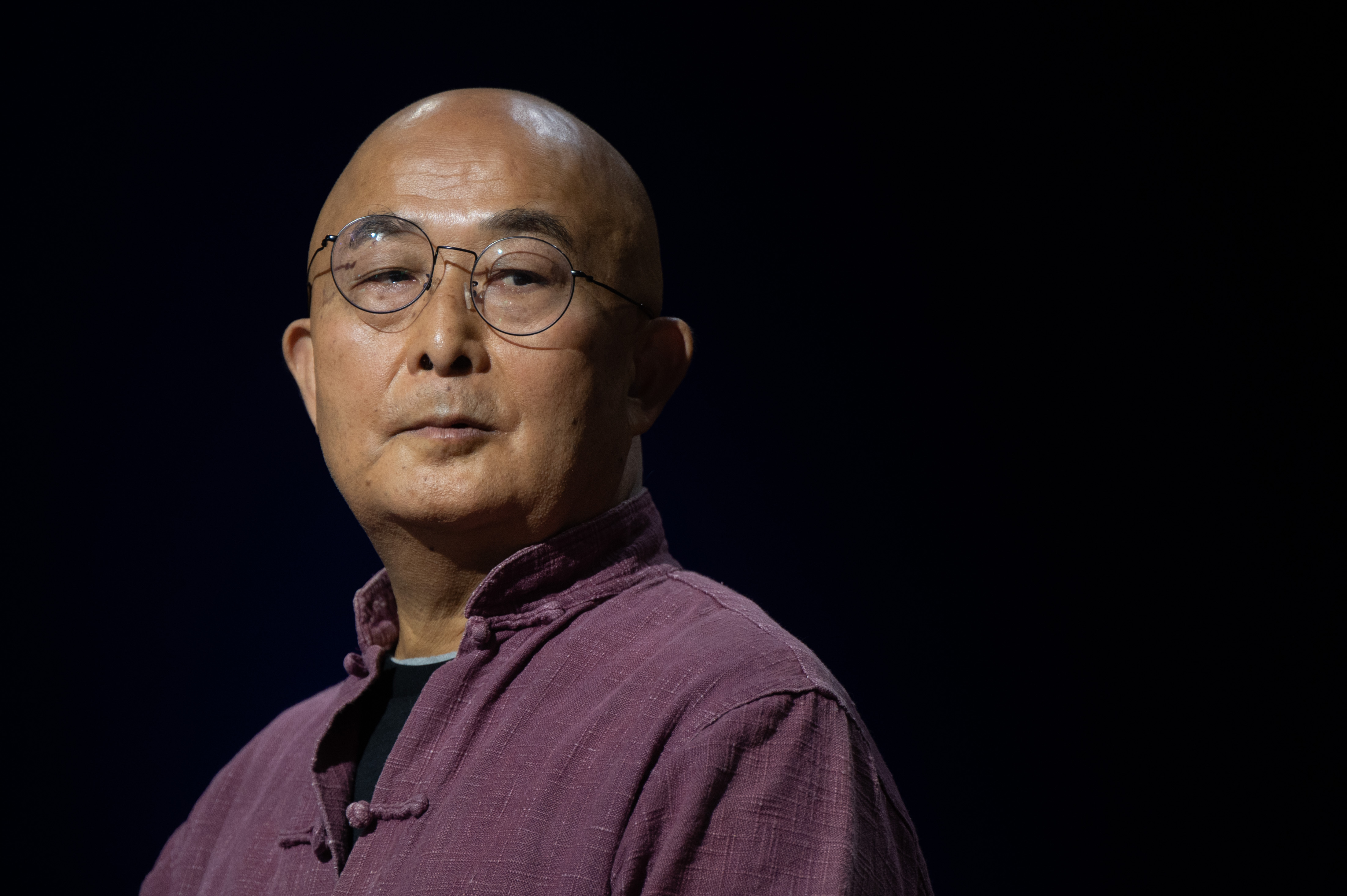 Der chinesische Schriftsteller Liao Yiwu blickt im lila Hemd mit hochgestelltem Kragen, mit Nickelbrille und kahlgeschorenem Kopf über seine Schulter.